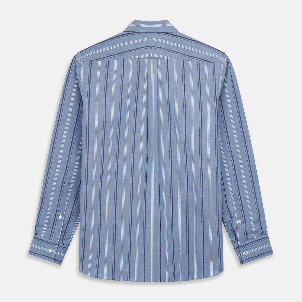 A man stands against a plain tan background wearing Turnbull & Asser's Blue Multi Chambray Stripe Weekend Fit Shirt with a Derby collar and 1 button cuffs, paired with a red patterned tie and gray trousers. His hands are in his pockets.