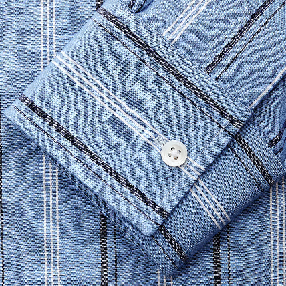 A man stands against a plain tan background wearing Turnbull & Asser's Blue Multi Chambray Stripe Weekend Fit Shirt with a Derby collar and 1 button cuffs, paired with a red patterned tie and gray trousers. His hands are in his pockets.