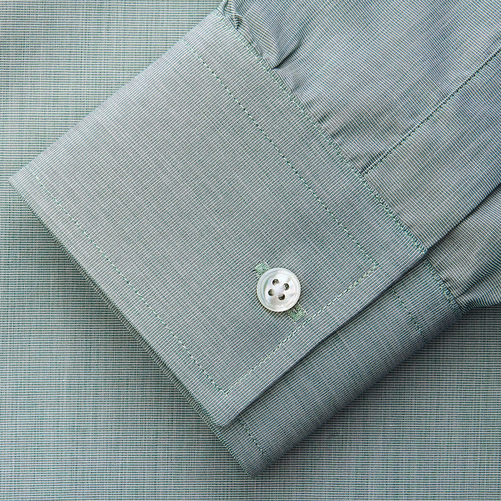 A man stands against a beige background wearing a Turnbull & Asser Green Weekend Fit Shirt with Long Point Collar and Single Button Cuffs, paired with a striped tie, dark trousers, and black dress shoes, hands in pockets and a neutral expression.