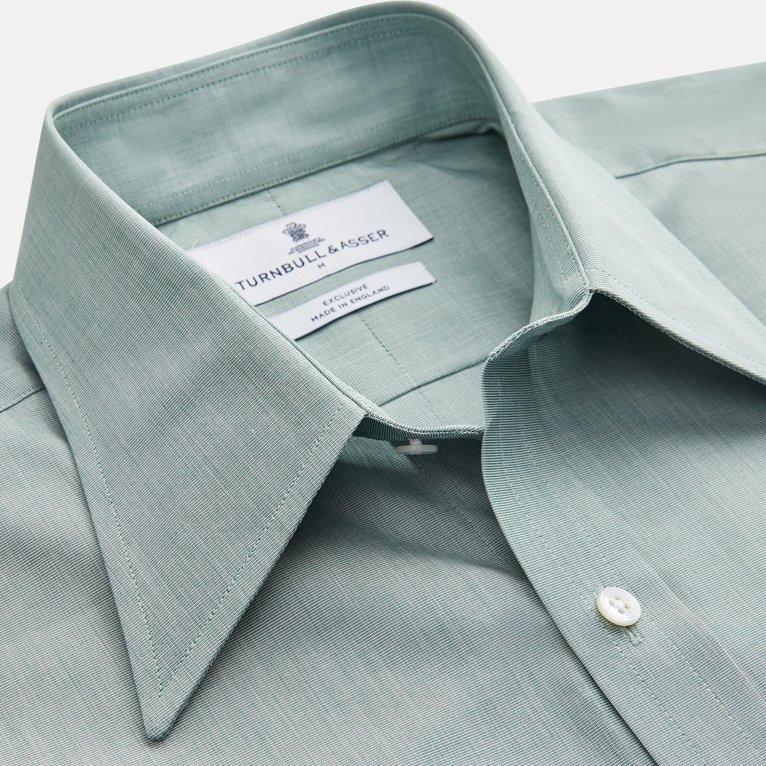View 3 of product A man stands against a beige background wearing a Turnbull & Asser Green Weekend Fit Shirt with Long Point Collar and Single Button Cuffs, paired with a striped tie, dark trousers, and black dress shoes, hands in pockets and a neutral expression.