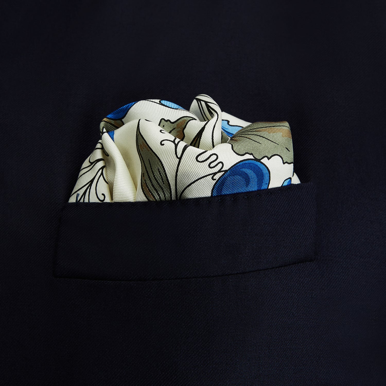 View 2 of product The Turnbull & Asser Pale Blue and Oat Foliage Pattern Silk Pocket Square is crafted in England, featuring a cream background with a circular blue and green floral design, bordered edges, and the brand's logo in one corner.