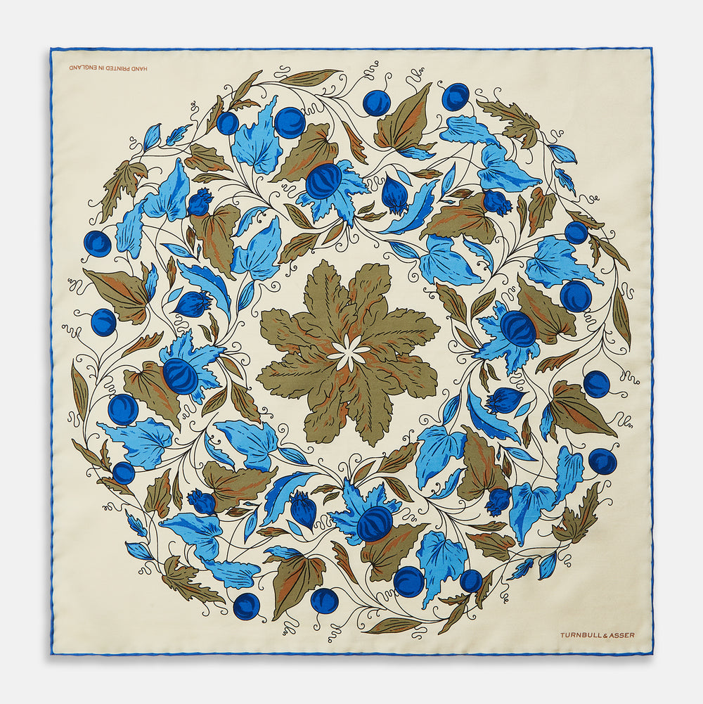 The Turnbull & Asser Pale Blue and Oat Foliage Pattern Silk Pocket Square is crafted in England, featuring a cream background with a circular blue and green floral design, bordered edges, and the brand's logo in one corner.