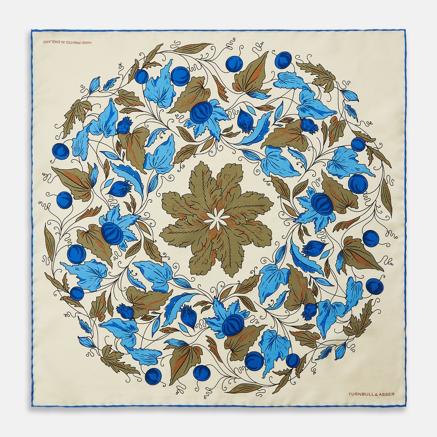 View 1 of product The Turnbull & Asser Pale Blue and Oat Foliage Pattern Silk Pocket Square is crafted in England, featuring a cream background with a circular blue and green floral design, bordered edges, and the brand's logo in one corner.