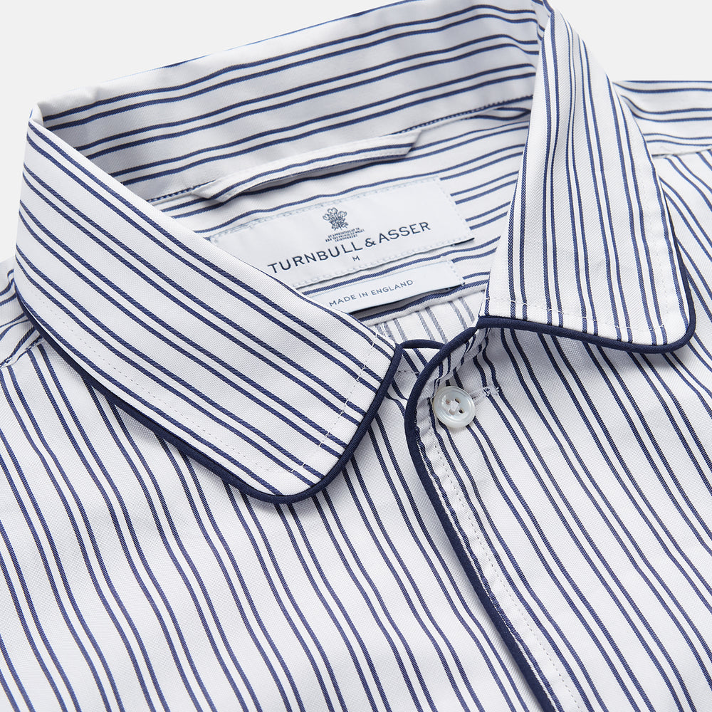The Turnbull & Asser White and Navy Striped Cotton Sussex Nightshirt features long sleeves, a knee-length cut, a collar, front buttons, a chest pocket with dark trim, and a curved hem for modern comfort inspired by tradition.