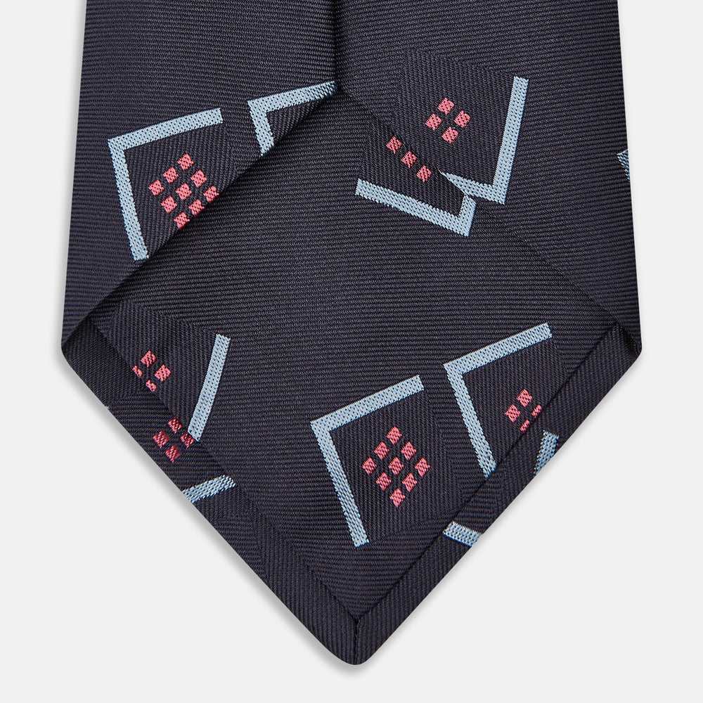 A Turnbull & Asser Navy Graphic Deco Silk Tie, featuring light blue chevrons and red squares on a dark navy background, shown rolled up against a plain white backdrop.