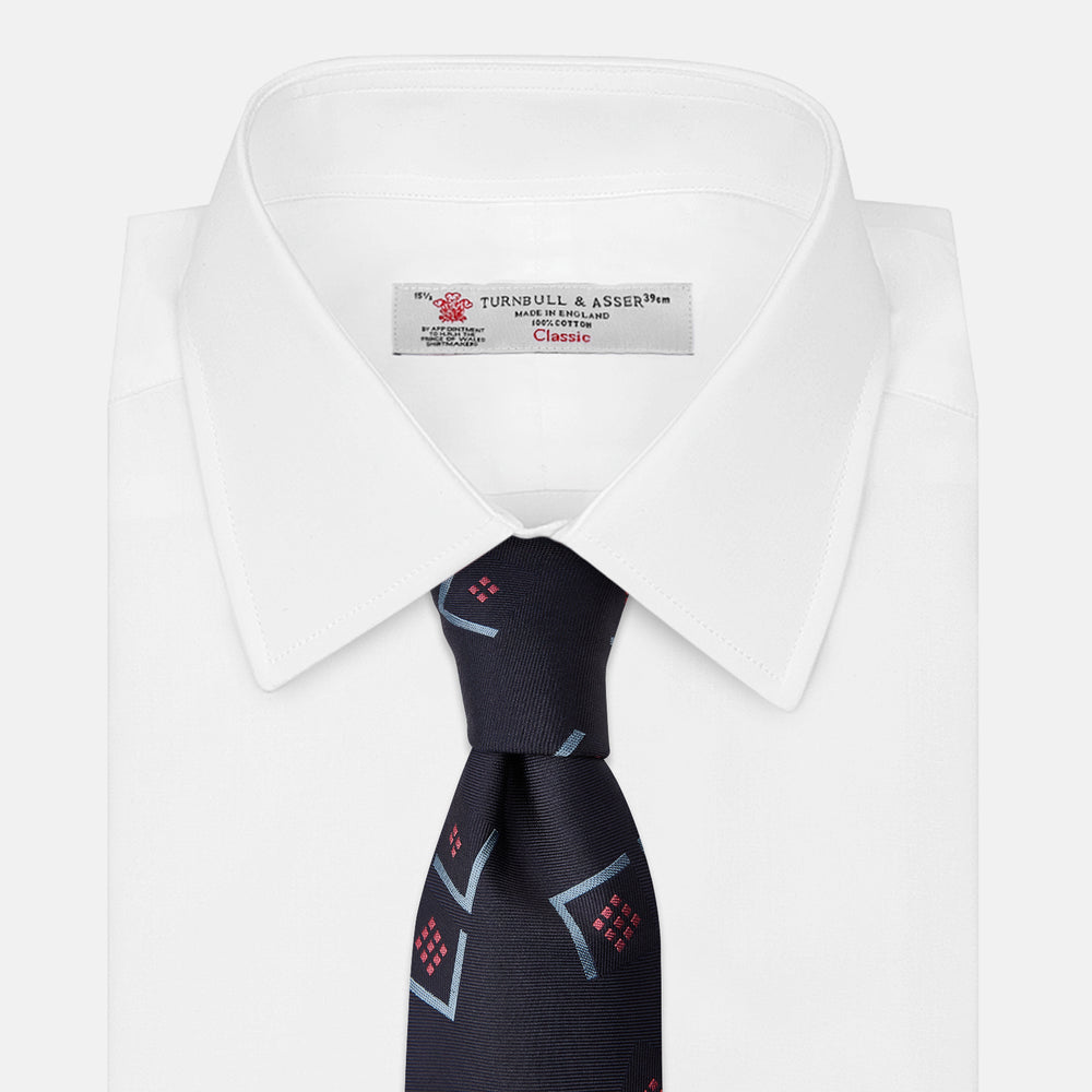 A Turnbull & Asser Navy Graphic Deco Silk Tie, featuring light blue chevrons and red squares on a dark navy background, shown rolled up against a plain white backdrop.
