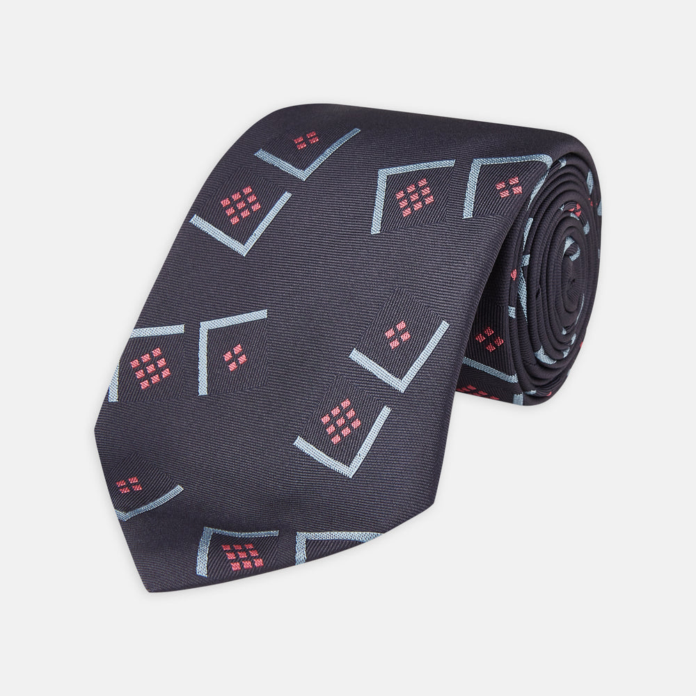 A Turnbull & Asser Navy Graphic Deco Silk Tie, featuring light blue chevrons and red squares on a dark navy background, shown rolled up against a plain white backdrop.