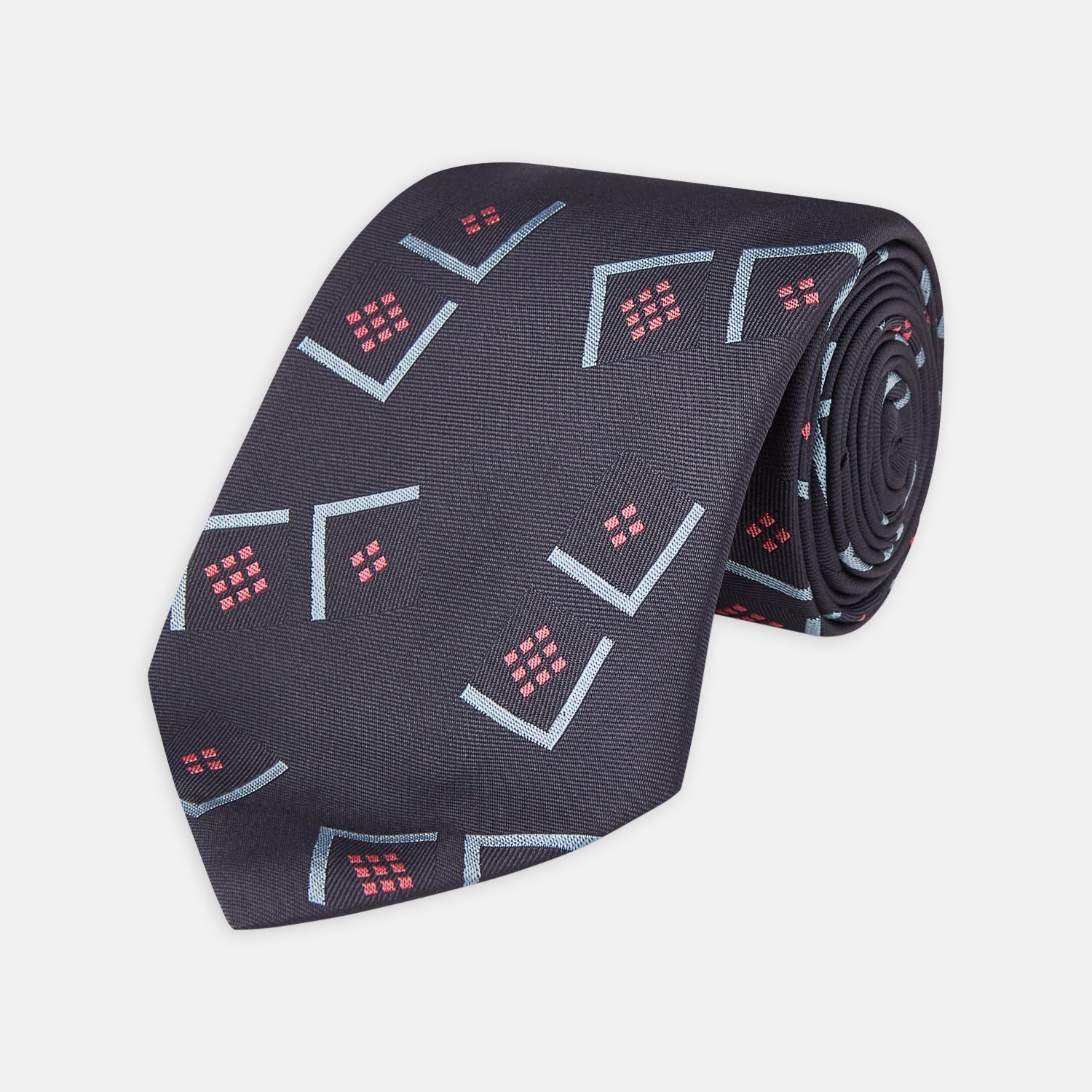 View 1 of product A Turnbull & Asser Navy Graphic Deco Silk Tie, featuring light blue chevrons and red squares on a dark navy background, shown rolled up against a plain white backdrop.