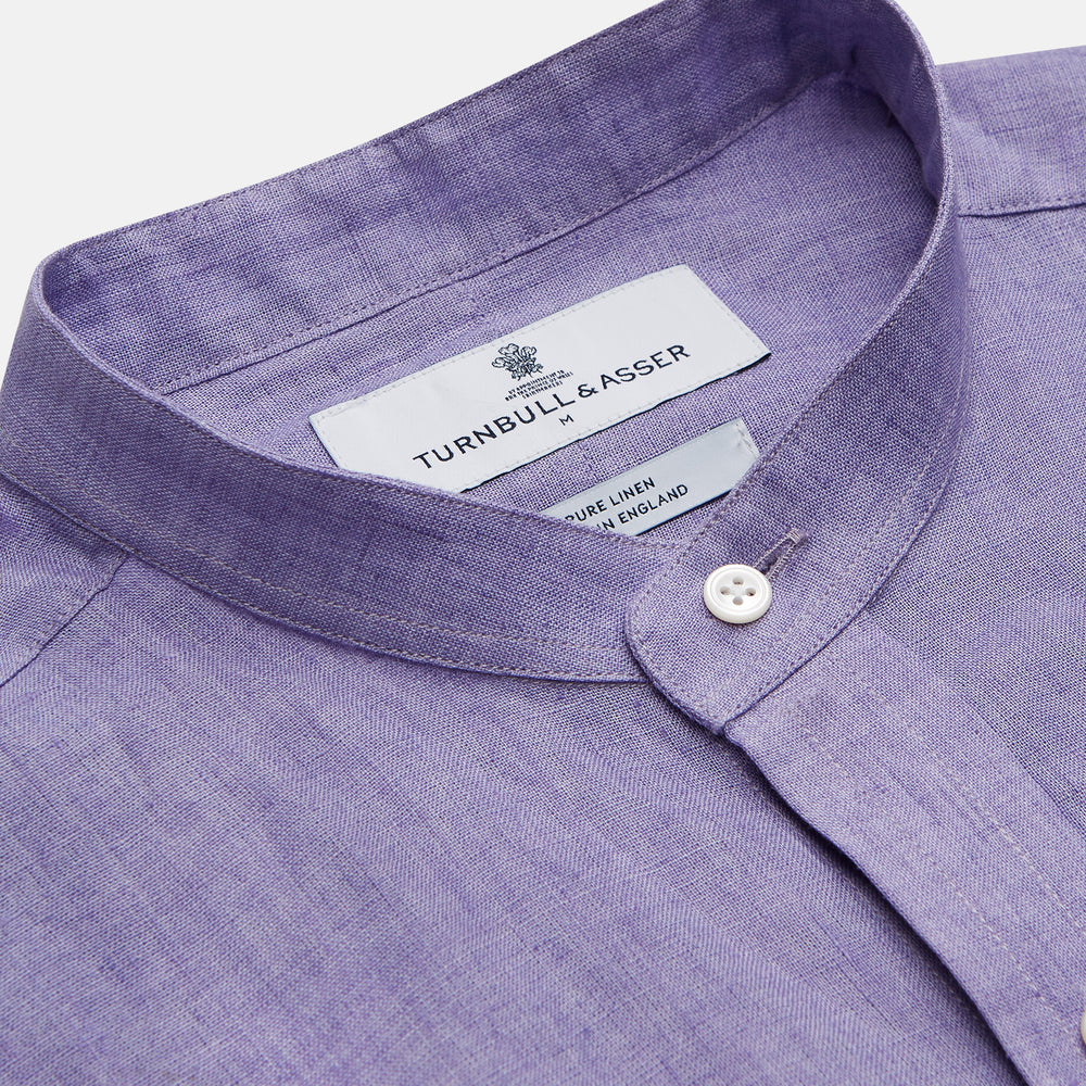 A man stands against a beige background wearing the Turnbull & Asser Lilac Weekend Fit Linen Blake Shirt with Stand Collar and 1-Button Cuffs, paired with dark pinstriped pants and brown sandals—a relaxed weekend look.