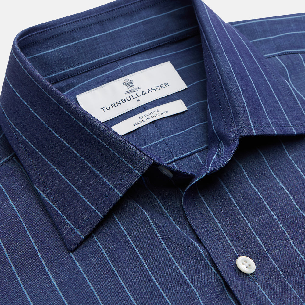 Close-up of a person wearing the Turnbull & Asser Blue Stripe Weekend Fit Nevis Shirt with Derby Collar and 1-Button Cuffs under a dark pinstripe blazer, focusing on the collar and upper chest. The face is partially out of frame.
