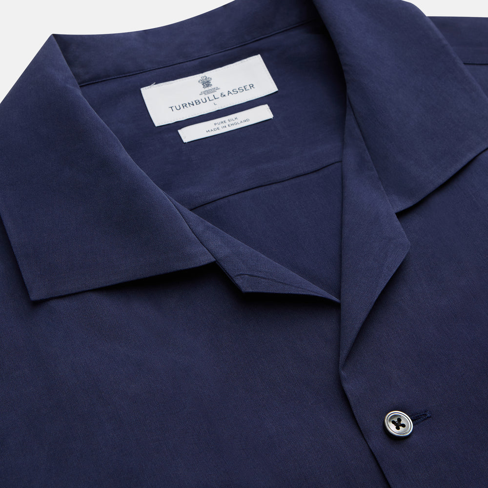 A man stands against a beige backdrop, wearing the Turnbull & Asser Navy Holiday Fit Silk Phillips Shirt with Revere Collar and dark pinstripe pants. He gazes downwards with a relaxed expression, embodying effortless summer style.