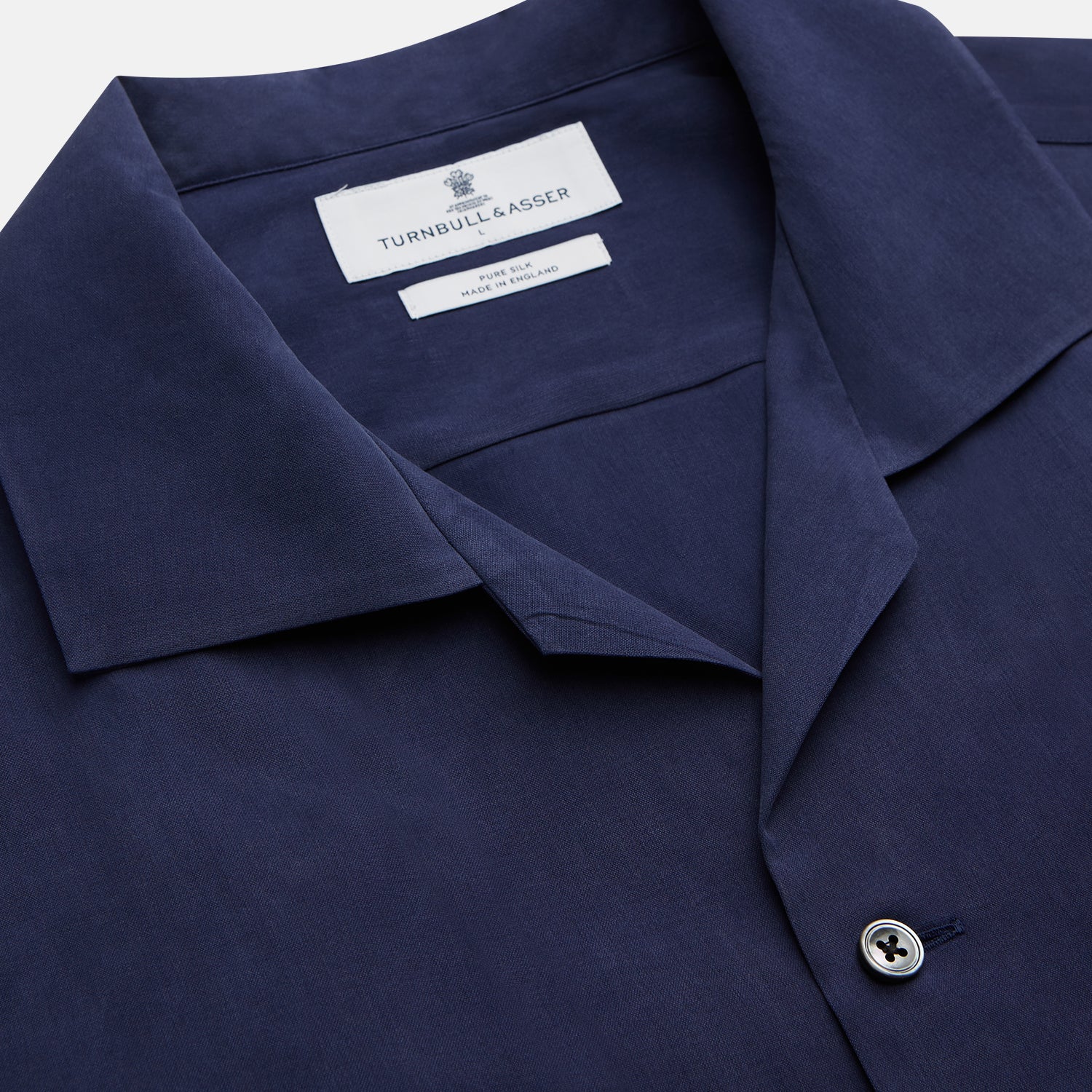 View 3 of product A man stands against a beige backdrop, wearing the Turnbull & Asser Navy Holiday Fit Silk Phillips Shirt with Revere Collar and dark pinstripe pants. He gazes downwards with a relaxed expression, embodying effortless summer style.