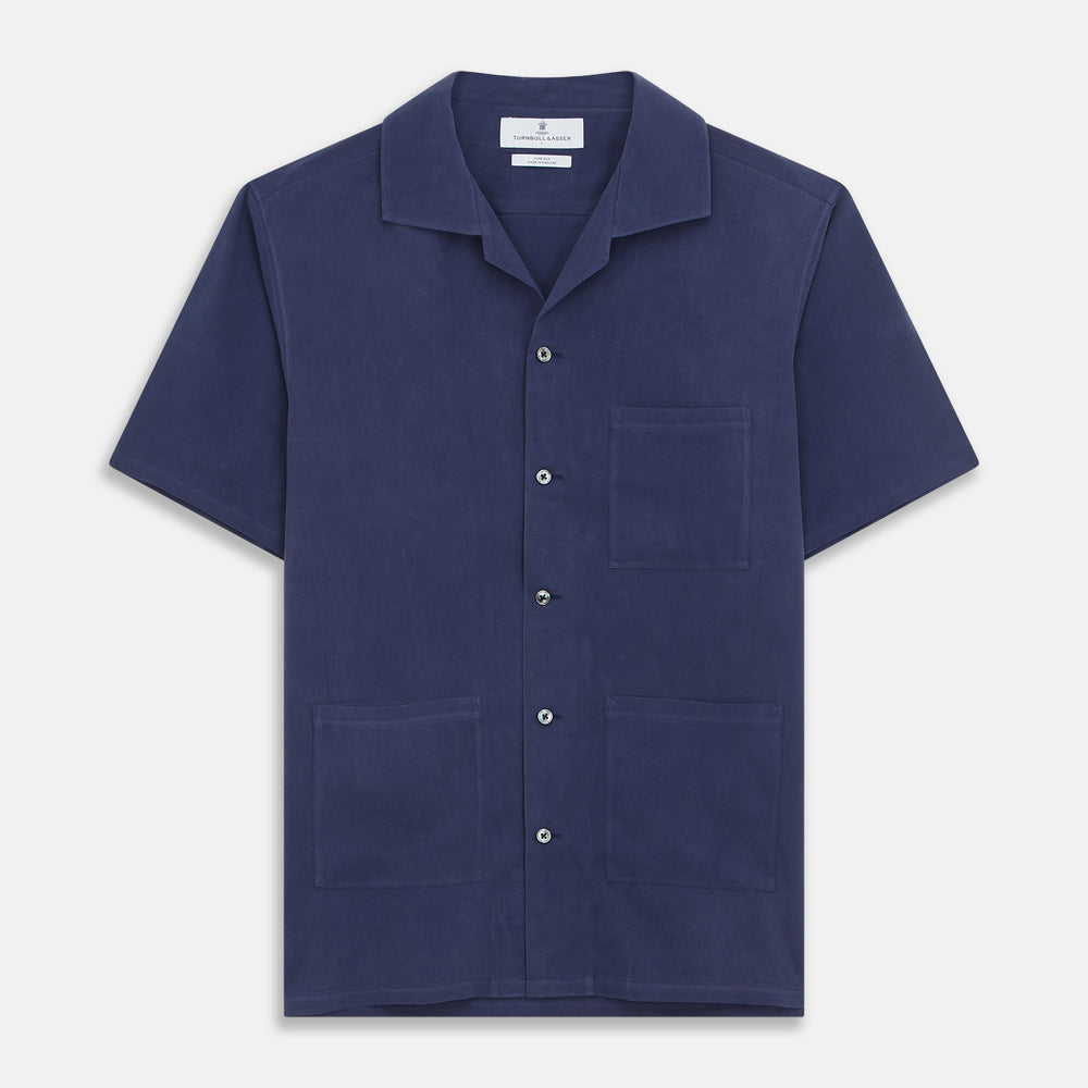 A man stands against a beige backdrop, wearing the Turnbull & Asser Navy Holiday Fit Silk Phillips Shirt with Revere Collar and dark pinstripe pants. He gazes downwards with a relaxed expression, embodying effortless summer style.