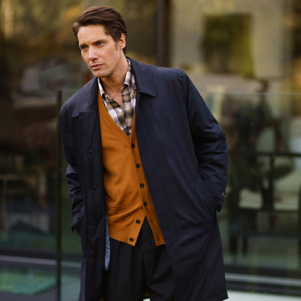 A man with brown hair stands outdoors, hands in his pockets, wearing a Turnbull & Asser Navy Sealup Quilted Liner Quintern Car Coat over a mustard cardigan. A glass barrier and blurred background are behind him.