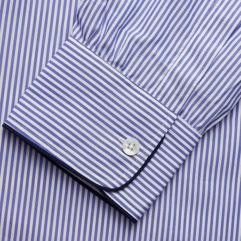 The Turnbull & Asser Blue Bengal Stripe Piped Cotton Nightshirt features long sleeves, a pointed collar, dark-trimmed pocket, curved hem, button details, and cuffed sleeves for an elegant look.