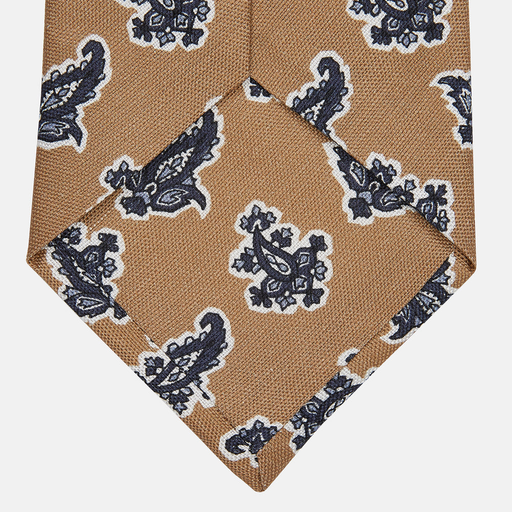 The Turnbull & Asser Toffee Paisley Floral Cotton Silk Blend Tie features a rolled-up design with dark blue and white paisley motifs on a tan background.