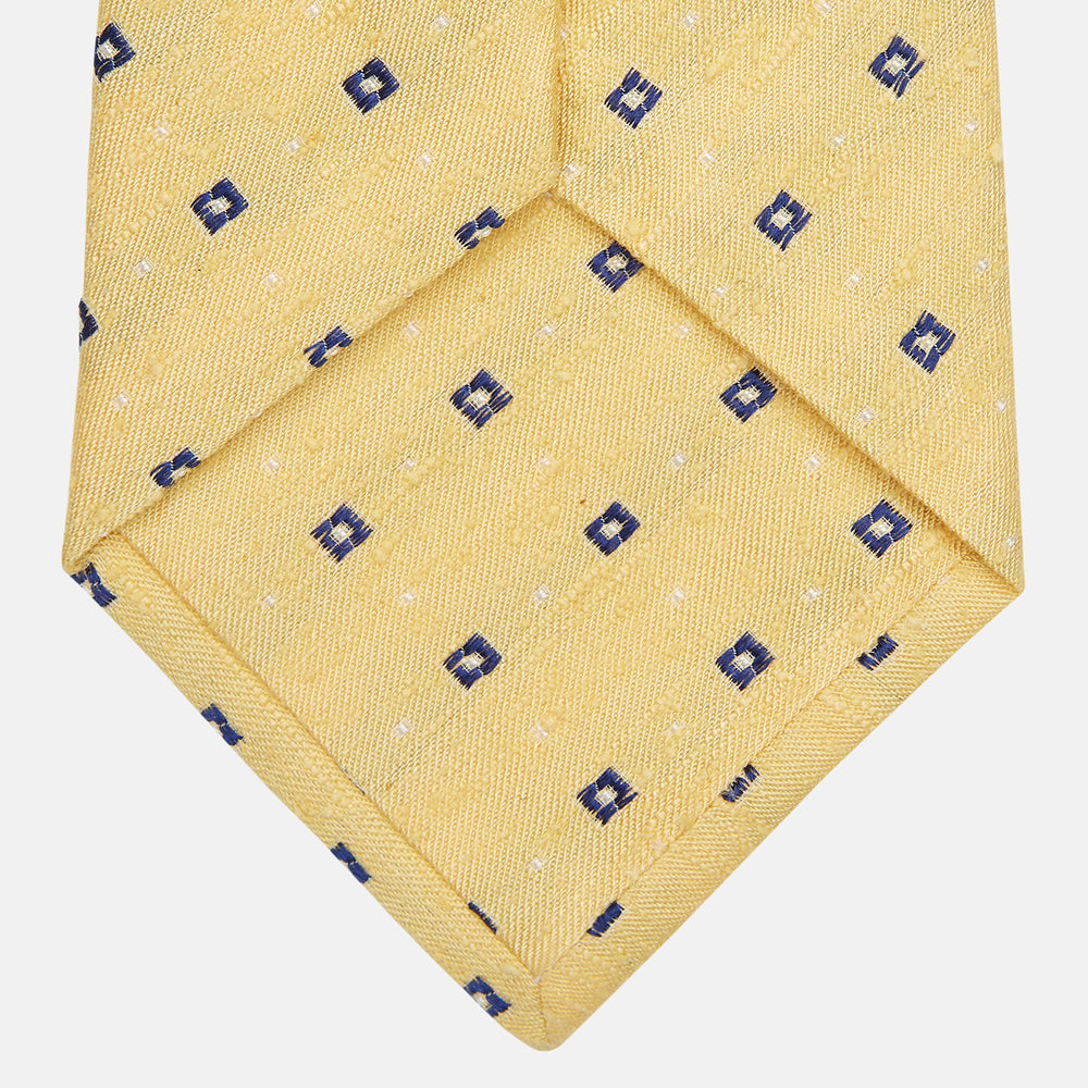 The Turnbull & Asser Yellow Fine Square Slub Silk Tie features a soft yellow jacquard with a subtle texture, decorated with a repeating blue and white diamond pattern, shown rolled up on a plain white background.