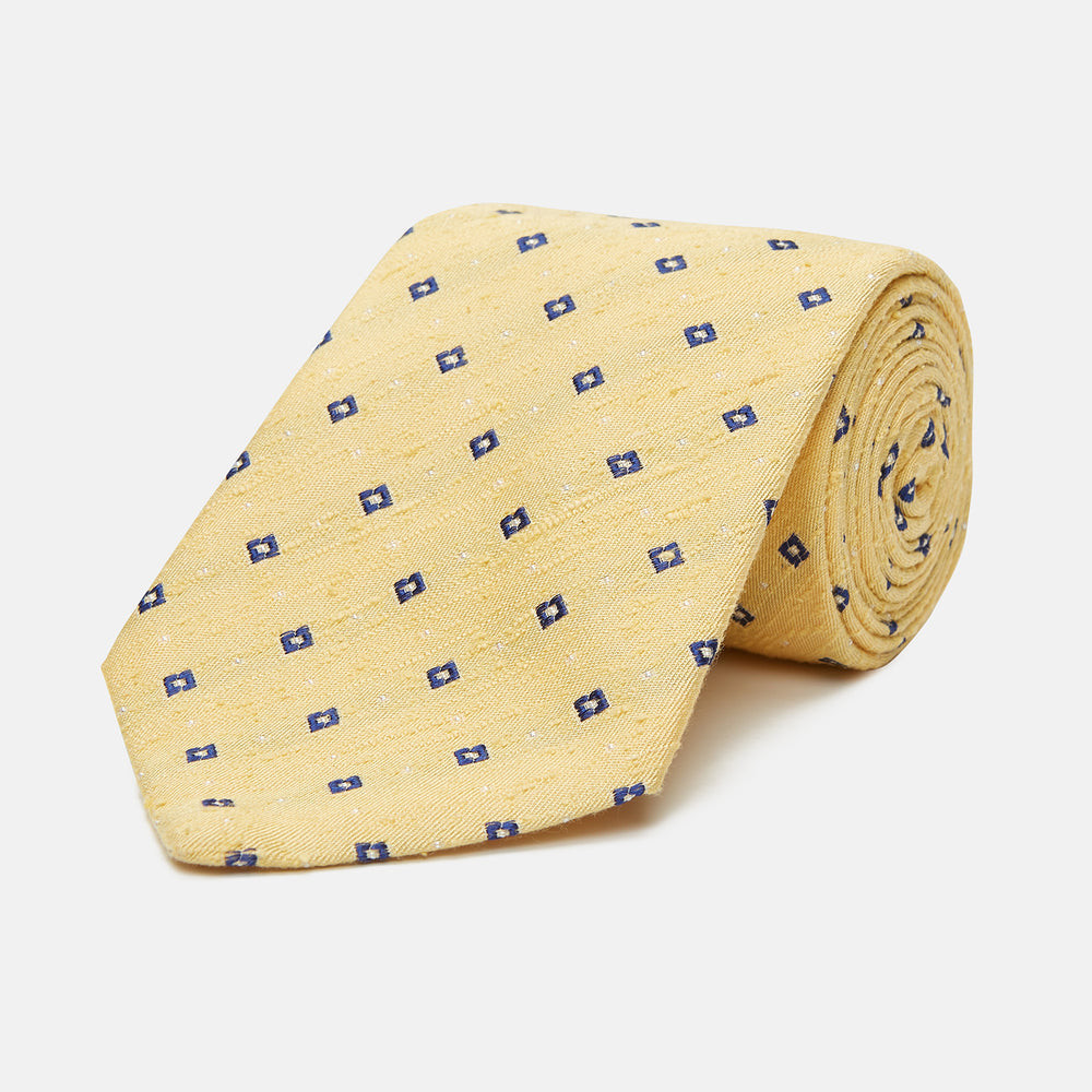 The Turnbull & Asser Yellow Fine Square Slub Silk Tie features a soft yellow jacquard with a subtle texture, decorated with a repeating blue and white diamond pattern, shown rolled up on a plain white background.