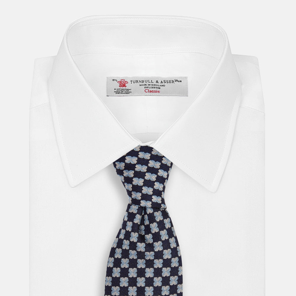 The Turnbull & Asser Indigo Graphic Floral Silk Jacquard Tie features a repeating pattern of light blue and white geometric shapes, shown rolled up on a plain white background.