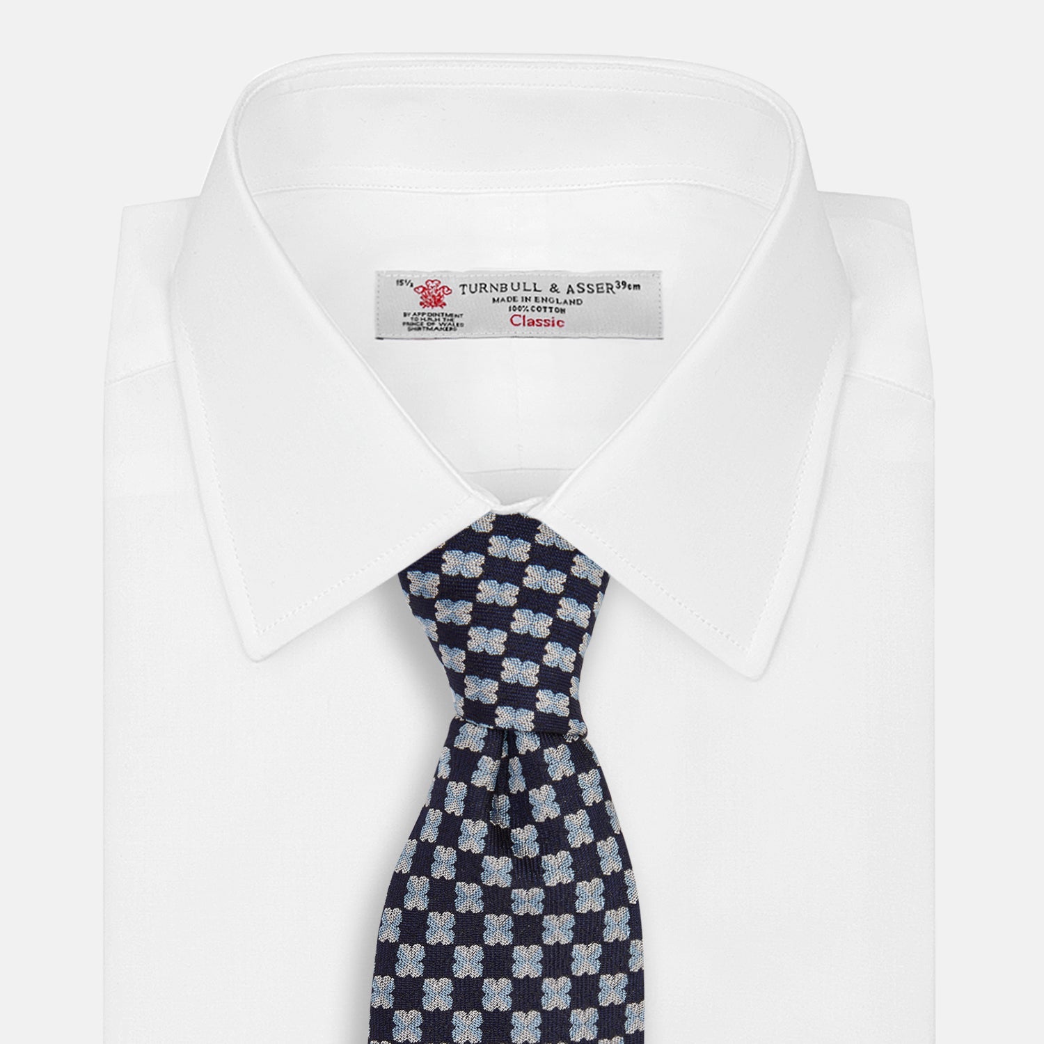 View 4 of product The Turnbull & Asser Indigo Graphic Floral Silk Jacquard Tie features a repeating pattern of light blue and white geometric shapes, shown rolled up on a plain white background.