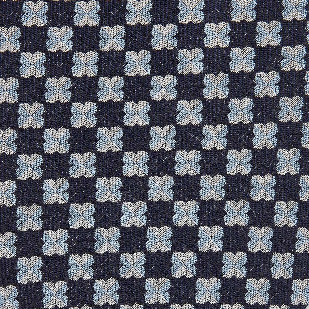 The Turnbull & Asser Indigo Graphic Floral Silk Jacquard Tie features a repeating pattern of light blue and white geometric shapes, shown rolled up on a plain white background.