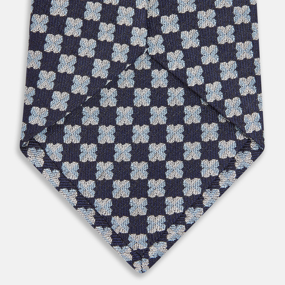 The Turnbull & Asser Indigo Graphic Floral Silk Jacquard Tie features a repeating pattern of light blue and white geometric shapes, shown rolled up on a plain white background.