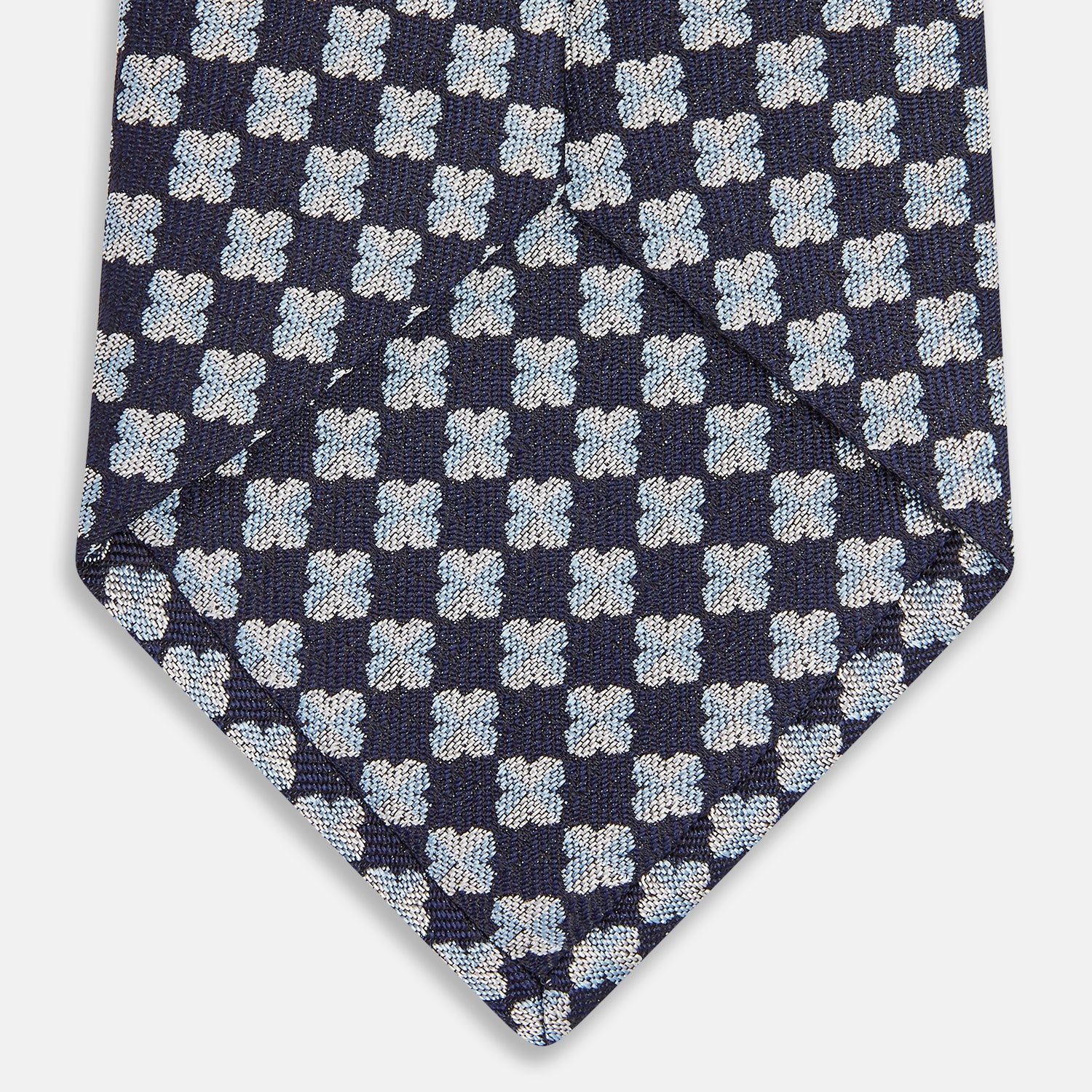 View 3 of product The Turnbull & Asser Indigo Graphic Floral Silk Jacquard Tie features a repeating pattern of light blue and white geometric shapes, shown rolled up on a plain white background.