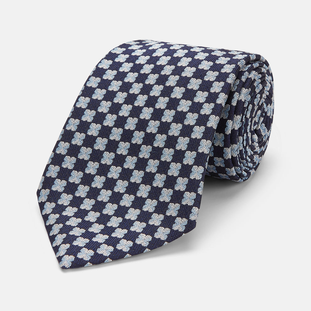 The Turnbull & Asser Indigo Graphic Floral Silk Jacquard Tie features a repeating pattern of light blue and white geometric shapes, shown rolled up on a plain white background.