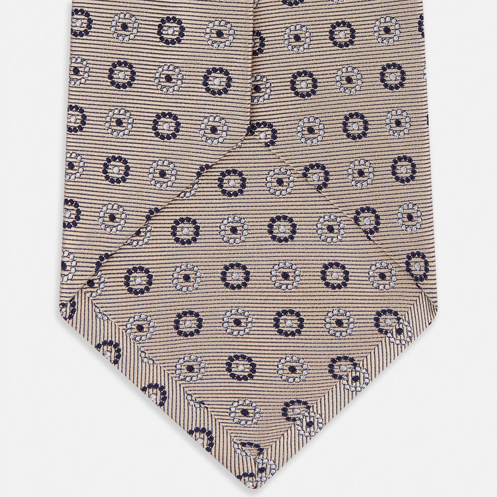 The Turnbull & Asser Beige Spheric Emblem Tie, a formalwear essential, features a neat roll and a repeating pattern of navy and white circular floral motifs on pastel beige.