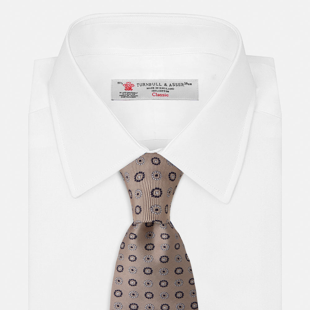 The Turnbull & Asser Beige Spheric Emblem Tie, a formalwear essential, features a neat roll and a repeating pattern of navy and white circular floral motifs on pastel beige.