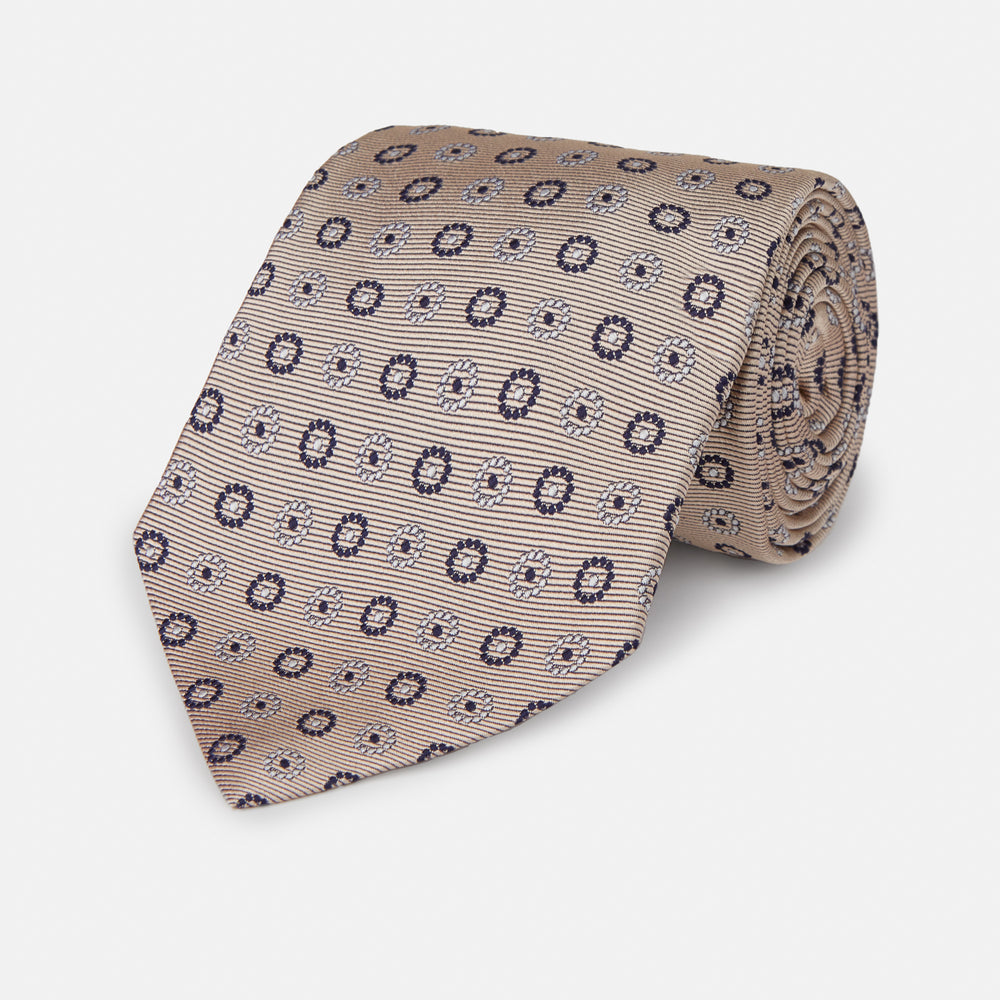 The Turnbull & Asser Beige Spheric Emblem Tie, a formalwear essential, features a neat roll and a repeating pattern of navy and white circular floral motifs on pastel beige.