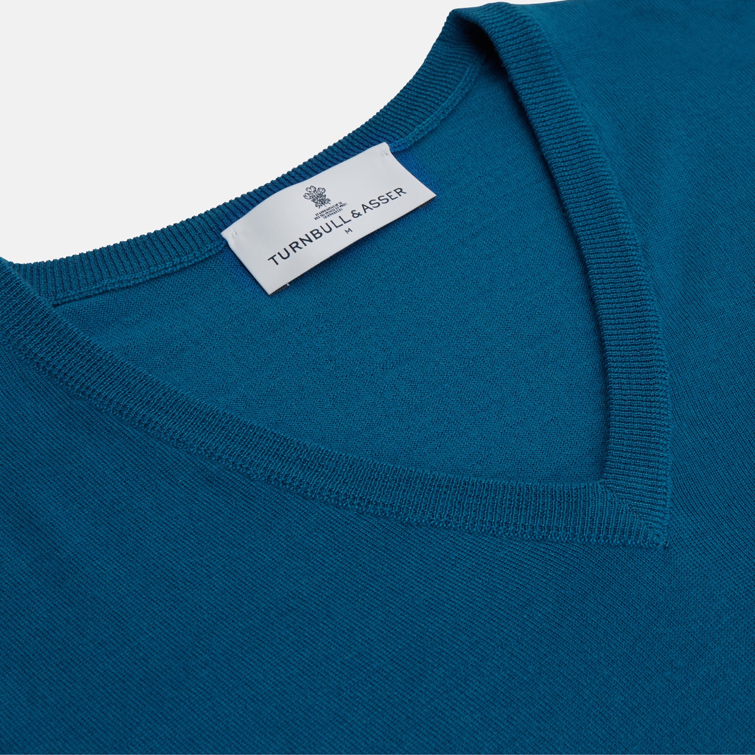View 2 of product The Turnbull & Asser Ocean Blue Merino Kennedy V-Neck Jumper, shown flat on a white background, features ribbed cuffs and hem with a visible branded label at the neckline.