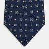 View 4 of product The Turnbull & Asser Navy Floral Emblem Tie displays a repeating pattern of small light blue, white, and yellow floral and diamond motifs, highlighting refined elegance, shown rolled up on a white background.