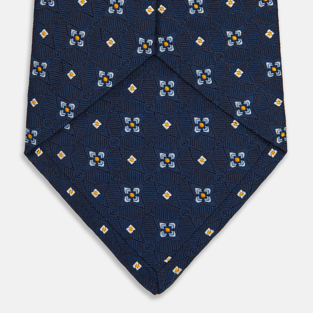 The Turnbull & Asser Navy Floral Emblem Tie displays a repeating pattern of small light blue, white, and yellow floral and diamond motifs, highlighting refined elegance, shown rolled up on a white background.