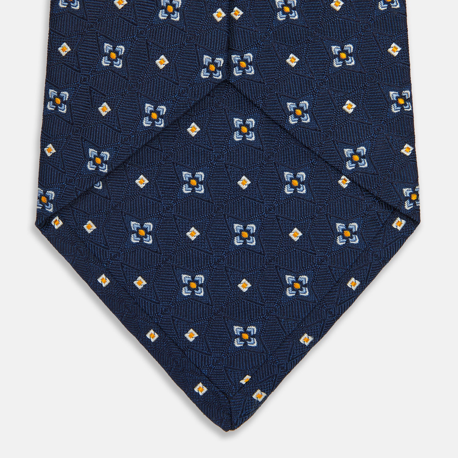 View 4 of product The Turnbull & Asser Navy Floral Emblem Tie displays a repeating pattern of small light blue, white, and yellow floral and diamond motifs, highlighting refined elegance, shown rolled up on a white background.