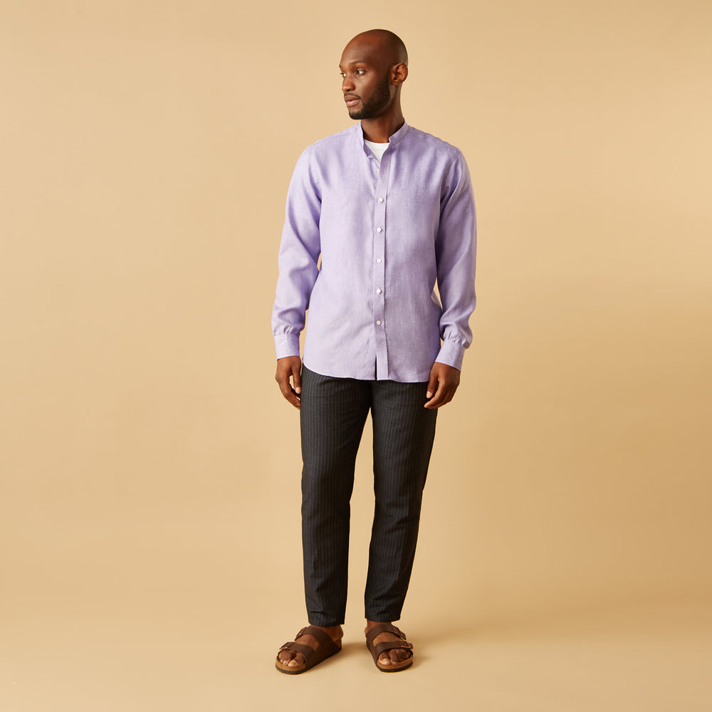 A man stands against a beige background wearing the Turnbull & Asser Lilac Weekend Fit Linen Blake Shirt with Stand Collar and 1-Button Cuffs, paired with dark pinstriped pants and brown sandals—a relaxed weekend look.