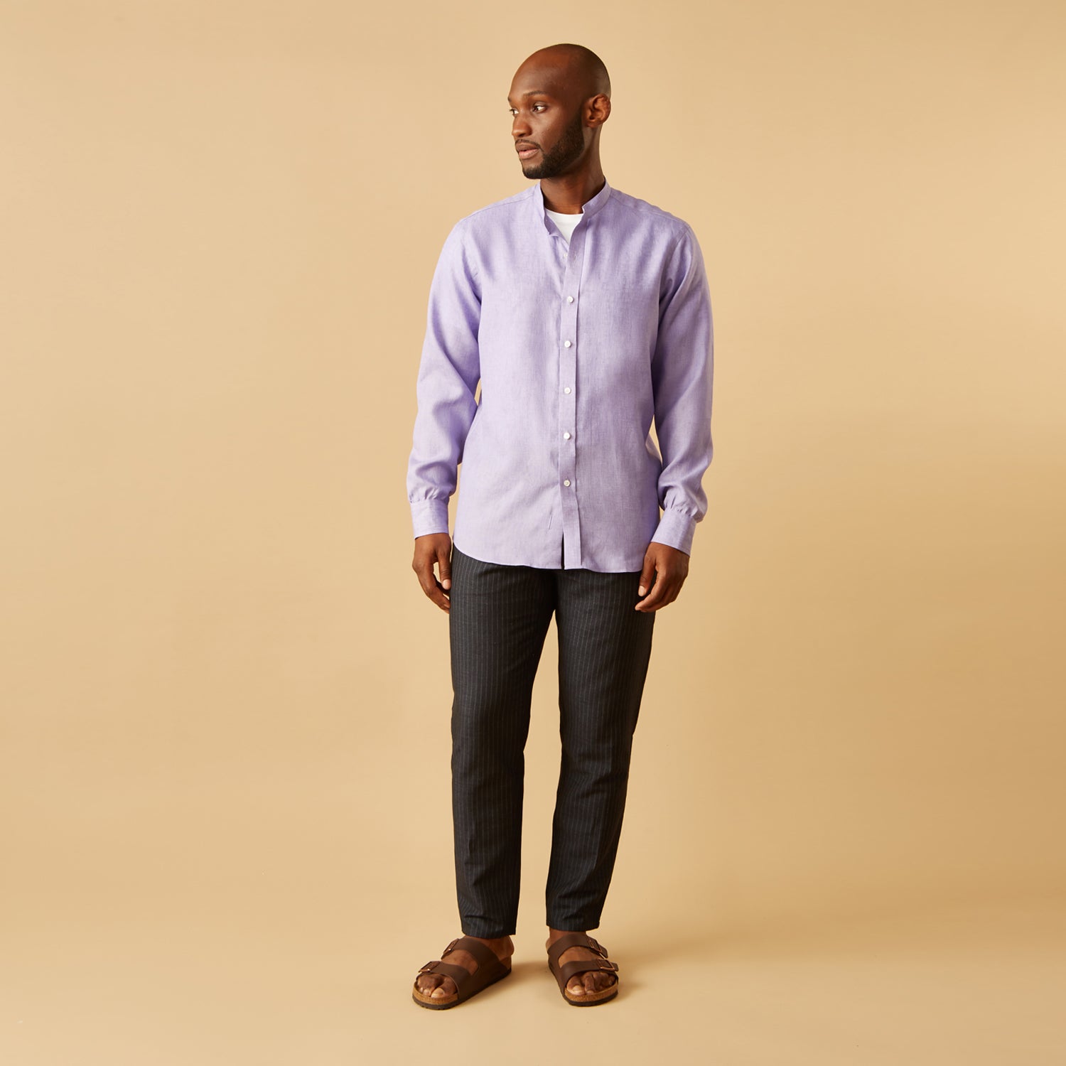 View 1 of product A man stands against a beige background wearing the Turnbull & Asser Lilac Weekend Fit Linen Blake Shirt with Stand Collar and 1-Button Cuffs, paired with dark pinstriped pants and brown sandals—a relaxed weekend look.