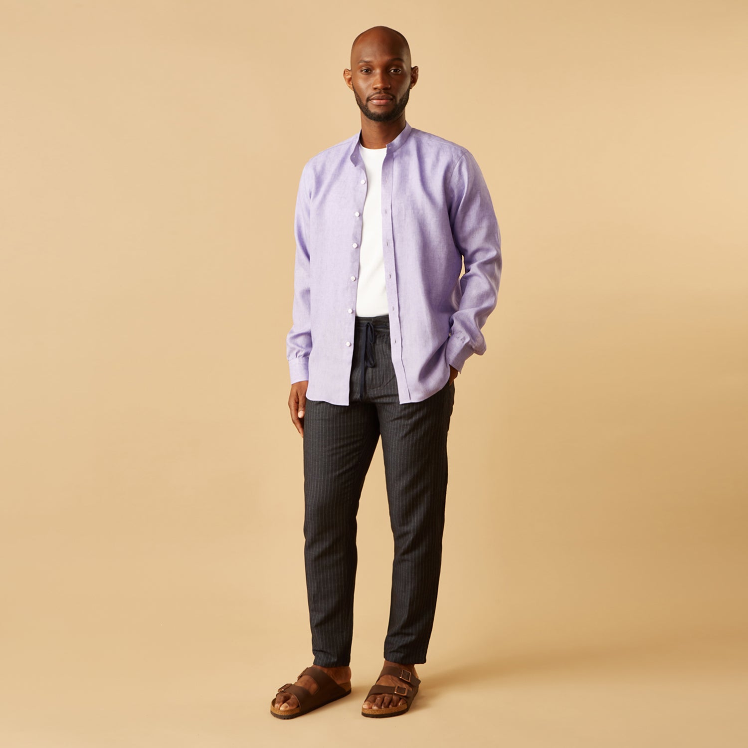 View 2 of product A man stands against a beige background wearing the Turnbull & Asser Lilac Weekend Fit Linen Blake Shirt with Stand Collar and 1-Button Cuffs, paired with dark pinstriped pants and brown sandals—a relaxed weekend look.