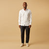 View 6 of product A man stands against a beige background, wearing the Turnbull & Asser White Linen Weekend Fit Blake Shirt, dark trousers, and beige sneakers. With one hand in his pocket and a neutral expression, he captures effortless weekend style.