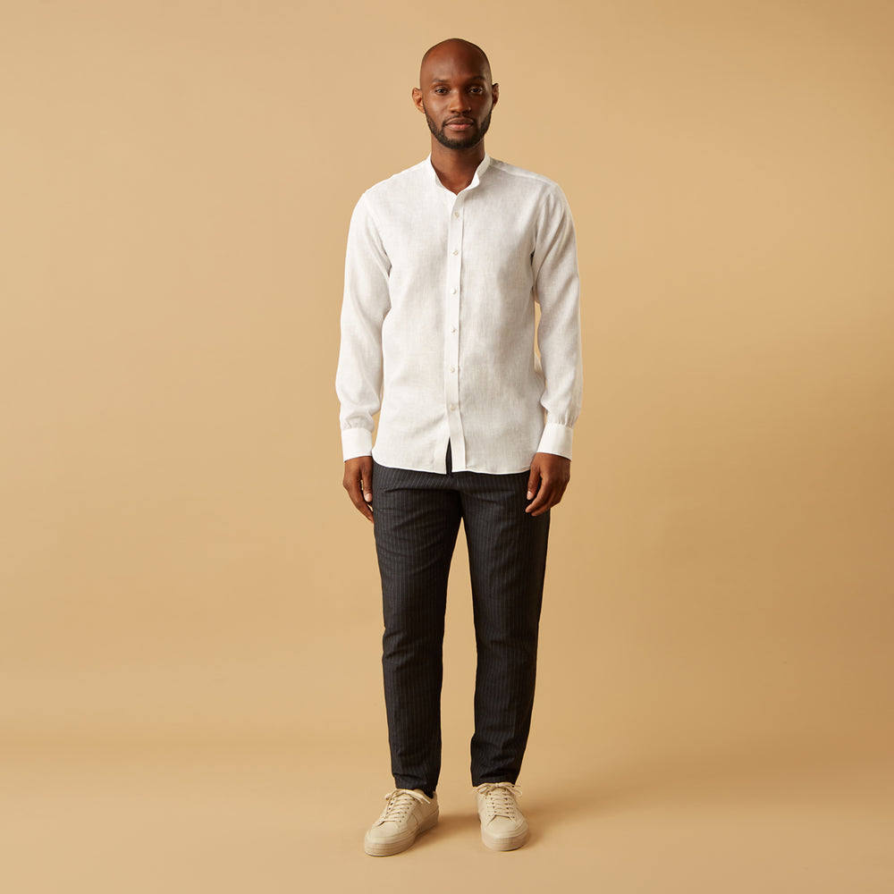 A man stands against a beige background, wearing the Turnbull & Asser White Linen Weekend Fit Blake Shirt, dark trousers, and beige sneakers. With one hand in his pocket and a neutral expression, he captures effortless weekend style.