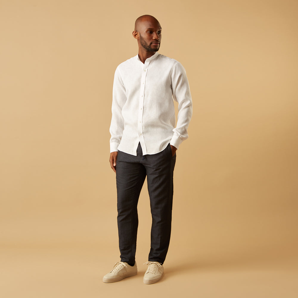A man stands against a beige background, wearing the Turnbull & Asser White Linen Weekend Fit Blake Shirt, dark trousers, and beige sneakers. With one hand in his pocket and a neutral expression, he captures effortless weekend style.