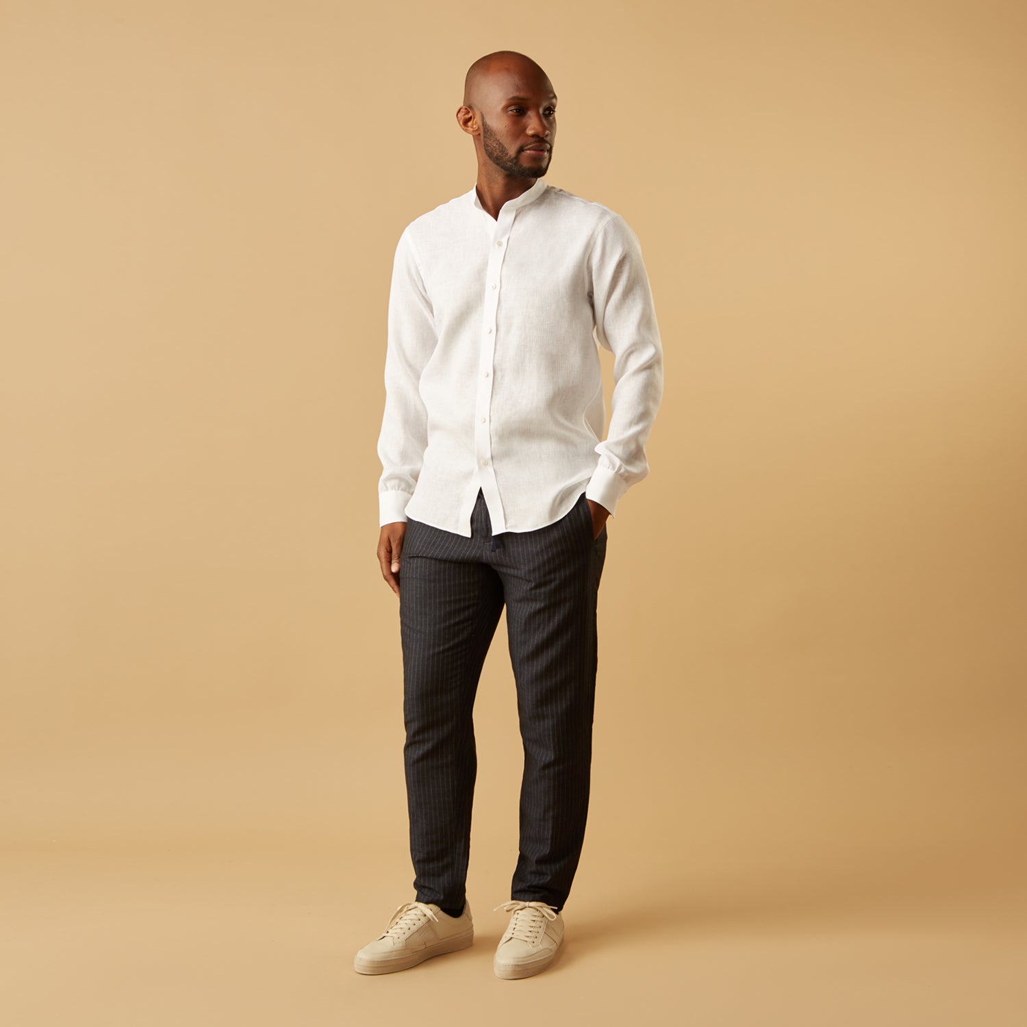 View 1 of product A man stands against a beige background, wearing the Turnbull & Asser White Linen Weekend Fit Blake Shirt, dark trousers, and beige sneakers. With one hand in his pocket and a neutral expression, he captures effortless weekend style.