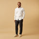 A man stands against a beige background, wearing the Turnbull & Asser White Linen Weekend Fit Blake Shirt, dark trousers, and beige sneakers. With one hand in his pocket and a neutral expression, he captures effortless weekend style.