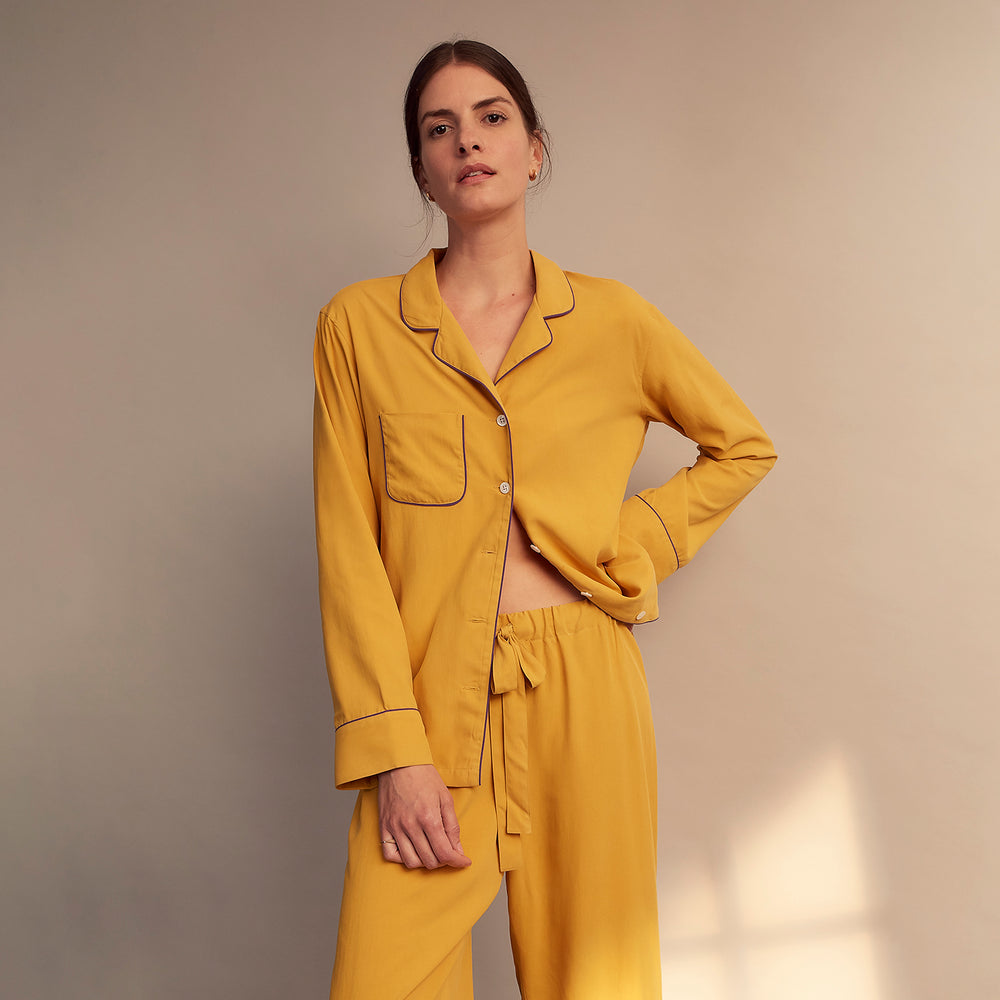 A woman stands barefoot on a gray floor wearing the Turnbull & Asser Women's Gold Silk Harriet Pyjamas Set, featuring a loose-fitting button-up top and long pants, against a plain beige background with soft light.