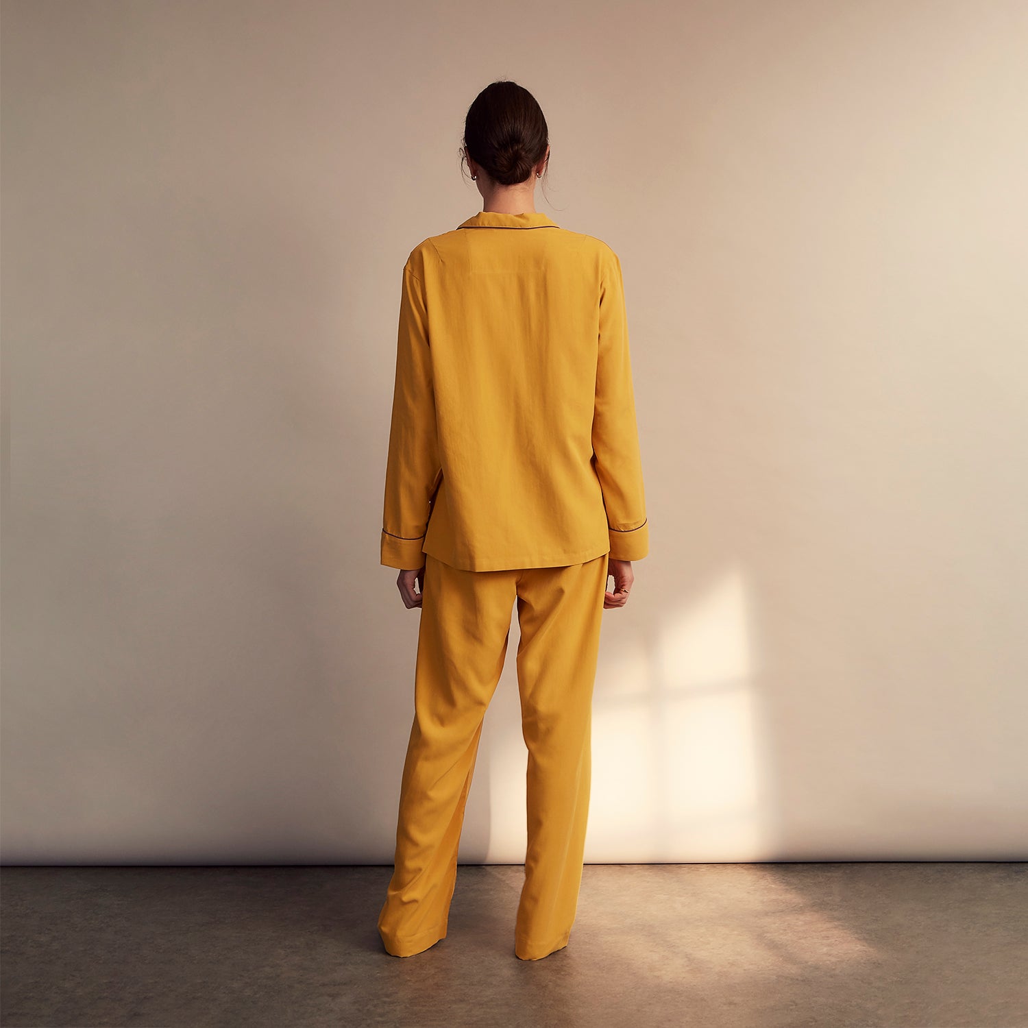 View 4 of product A woman stands barefoot on a gray floor wearing the Turnbull & Asser Women's Gold Silk Harriet Pyjamas Set, featuring a loose-fitting button-up top and long pants, against a plain beige background with soft light.