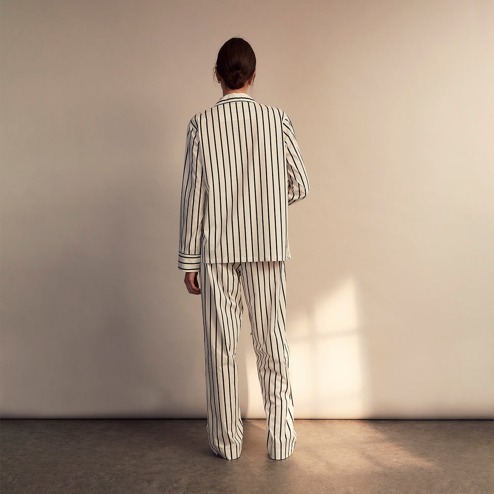 A person stands barefoot on a concrete floor, wearing the Turnbull & Asser Black & White Stripe Harriet Women’s Pyjama Set, featuring loose-fitting white pajamas with black vertical stripes, against a plain beige wall in soft side lighting.