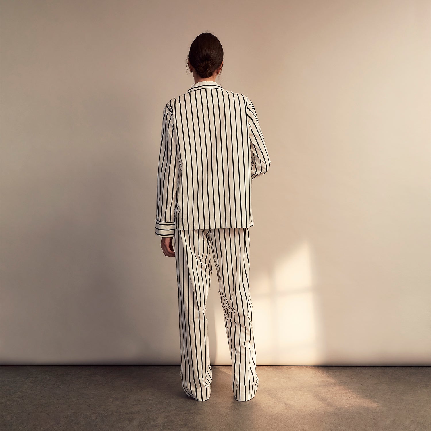 View 8 of product A person stands barefoot on a concrete floor, wearing the Turnbull & Asser Black & White Stripe Harriet Women’s Pyjama Set, featuring loose-fitting white pajamas with black vertical stripes, against a plain beige wall in soft side lighting.