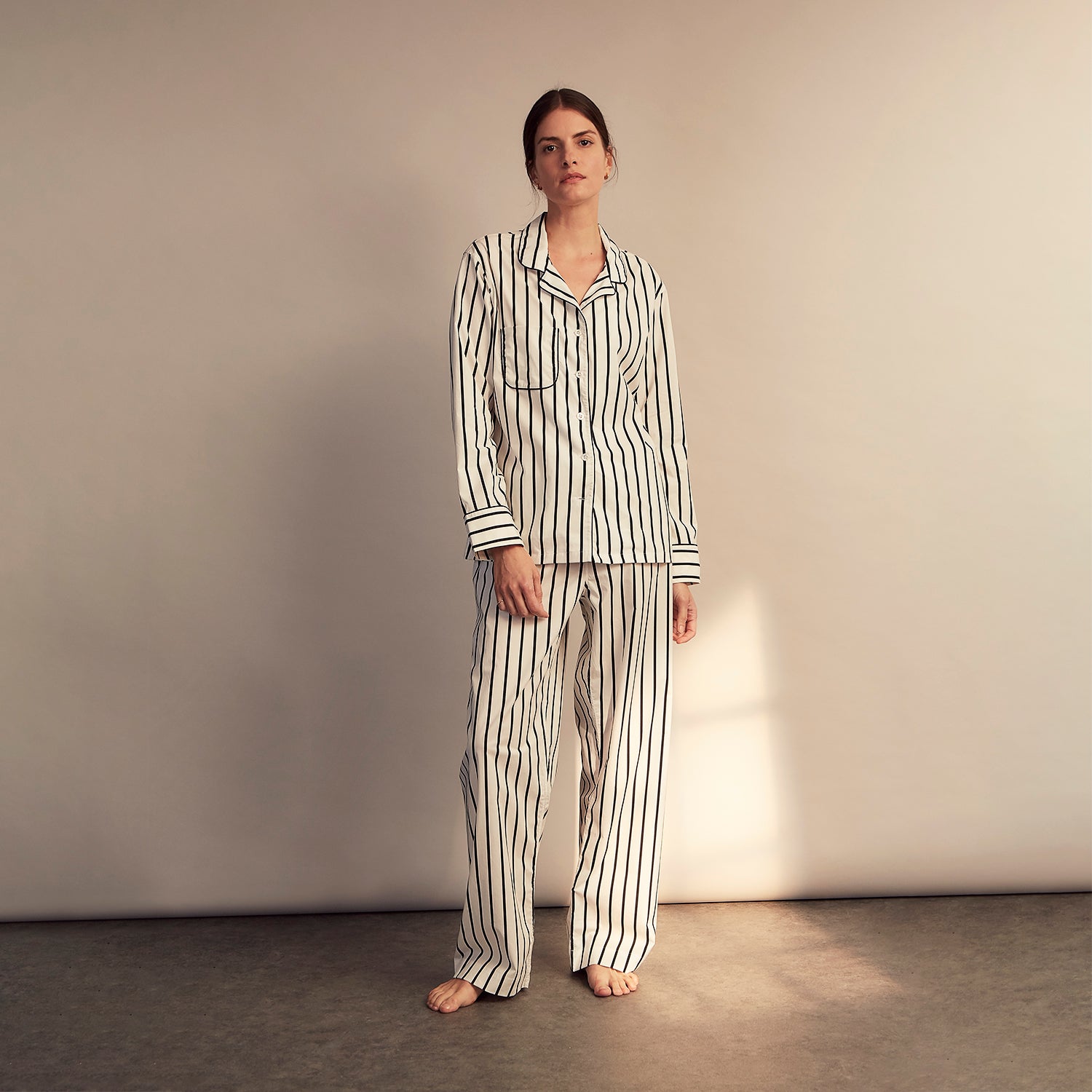 View 1 of product A person stands barefoot on a concrete floor, wearing the Turnbull & Asser Black & White Stripe Harriet Women’s Pyjama Set, featuring loose-fitting white pajamas with black vertical stripes, against a plain beige wall in soft side lighting.