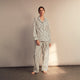 A person stands barefoot on a concrete floor, wearing the Turnbull & Asser Black & White Stripe Harriet Women’s Pyjama Set, featuring loose-fitting white pajamas with black vertical stripes, against a plain beige wall in soft side lighting.