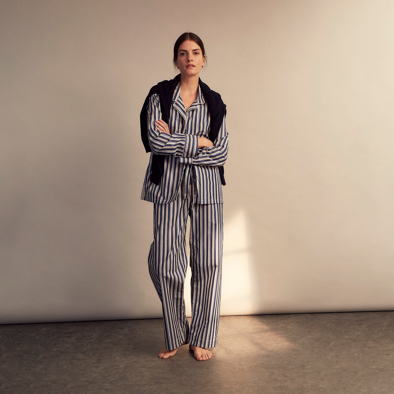 View 1 of product A person stands barefoot on a gray floor in the Turnbull & Asser Blue Multi Stripe Harriet Women’s Pyjama Set with a dark sweater over their shoulders—premium cotton pyjamas that make a perfect gift for her—against a plain beige background.